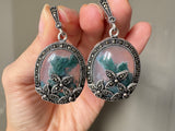 These one of a kind silver earrings feature oval dangles measuring 2 inches in length and adorned with delicate flower marcasite. The focal point of these earrings is the oval moss agate gemstone in a stunning shade of green. Exuding femininity and classic style, these earrings make for a special and unique addition to any jewelry collection, especially for grandmothers. These feminine and classic silver jewelry earrings are truly one of a kind, featuring oval dangle designs measuring 2 inches in length.