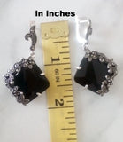 Black square earrings with silver detailing next to a ruler showing their size.