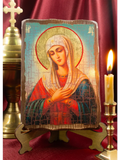 A handcrafted "Tenderness" Mother of God wooden icon displayed on a rustic stand next to a lit candle. The Virgin Mary is depicted with crossed arms in a moment of prayer, radiating a serene gold halo layout.