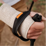 A close-up of a person outdoors wearing a beige wrist woolen wrist wrap with a black strap and small orange accent, holding a trekking pole. 