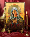 An Orthodox Christian icon of the Virgin Mary on a wooden stand, displayed on a red satin fabric next to a lit candle and a brass vessel. Close-up view of Virgin Mary icon with its signature humble expression and folded hands. This high-quality wooden religious icon shows rich green and gold Byzantine color choices.