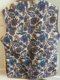 A sleeveless quilted vest with an all-over floral and paisley pattern in blue, brown, and beige, hanging on a black hanger against a white paneled wall.