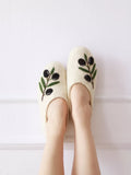 A person wearing cream-colored felt slippers with embroidered olive branches in black and green, standing against a white paneled wall.