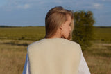 A woman standing in a grassy field, viewed from behind, wearing a cream-colored sleeveless warm woolen vest over a white dress. 