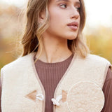 A woman wearing a cream-colored sheepskin vest over a brown ribbed sweater, with a yellow promotional text banner and a circular fabric detail inset. 
