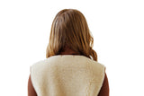 A woman shown from the back wearing a beige sherpa woolen vest over a brown long-sleeve top and light blue jeans.