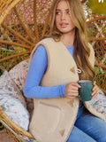 A woman sits in a woven hanging chair wearing a beige modern woolen vest over a blue long-sleeve top and blue jeans, holding a dark green mug.