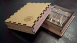 A light brown wooden Orthodox icon presentation box with interlocking puzzle-style edges, shown closed with a circular embossed logo on the lid, next to an open box containing a clear plastic-wrapped item, on a dark surface.