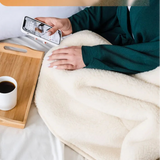 A person lying in bed under a soft cream sheep wool large blanket, holding a smartphone. A wooden tray with a cup of black coffee sits beside them, and an orange text banner appears at the top.