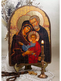 A traditional wooden Christian icon of the Holy Family (Mary, Joseph, and Jesus) displayed on a small wooden stand. The icon is styled with a lit candle in a brass holder, a wooden rosary, and a small wooden cross on a white lace doily, with dried flowers in the background. Handcrafted Holy Family wooden icon displayed on a mini table stand next to a burning wax candle. The varnished Christian artwork captures Jesus, Mary, and Joseph under golden halos.