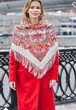 A woman looking away and smiling while wearing a heavy, traditional Slavic shawl draped over her shoulders. A high-quality ivory merino wool scarf featuring detailed paisley borders and dense floral bouquets worn over a red jacket.
