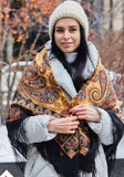 Woman wearing a patterned black, brown and orange woolen Slavic scarf and knit hat in an outdoor setting with trees and snow. A premium woolen palantine made from 100% merino wool, featuring a complex ethnic-inspired pattern in shades of orange, brown, and black, finished with soft fringes along the edges.