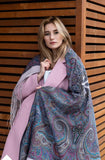 Woman wearing a pink coat and gray patterned merino wool very large scarf against a wooden background.  A warm winter palantine made of soft merino wool, showcasing an intricate Slavic folk pattern in shades of gray, ivory, and soft blue.