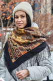 Woman wearing a patterned woolen triangle black and orange scarf and knit hat in a snowy outdoor setting. A large, square merino wool shawl styled as a palantine, draped elegantly to show its detailed woven pattern in deep black, vibrant orange, and rich brown with delicate fringe trim.