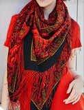 Red roses extra large flamenco red scarf with fringes worn by a person, against a neutral background. A model stands looking to the side, wrapped snugly in a large, colorful merino wool scarf designed in a traditional Slavic pattern. The thick, opaque red fabric is covered in vibrant rose arrangements and complex black border motifs, accentuating a classy and cozy winter fashion look. An outdoor lifestyle photograph of a woman modeling a premium red and black woolen shawl. 