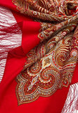 A product shot of a large, oversized merino wool shawl draped on white background. The shawl features a solid, vibrant red base that transitions into layers of intricate, traditional Slavic paisley patterns in brown, tan, and white. Long, matching red silk-like fringes hang down from the triangular hemline.