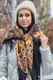Woman wearing a patterned scarf and knit hat in a forest setting. An elegant woolen shawl crafted from merino wool, showcasing a warm color palette of deep black, burnt orange, and earthy brown.