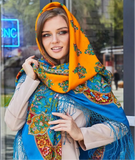 A woman wrapped snugly in a large, colorful merino wool palantine designed in a traditional Slavic pattern. The top layer creates a bright orange hood, while the main body of the blanket-style shawl cascades down in a vibrant blue print filled with detailed orange and red flowers, accentuating a cozy winter fashion look.