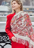 A woman stands outdoors wrapped snugly in a large, colorful merino wool scarf designed in a traditional Slavic pattern. The thick, opaque ivory fabric is covered in vibrant pink rose arrangements and complex border motifs, accentuating a classy and cozy winter fashion look.