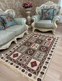 A rectangular area Armenian rug with a geometric tribal pattern in beige, brown, red, and black, styled on a light wood floor in a luxurious room with matching light blue sofas and ornate white armchairs.