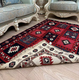 A traditional Armenian-style area rug with a red, beige, and navy geometric pattern, shown partially folded on a light wood floor in a living room setting with ornate light blue sofas.