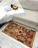 A rectangular woven area rug with a geometric Southwestern-style pattern in rust, terracotta, beige, and navy, styled on a light gray wood floor next to a cream-colored sofa with a striped throw blanket.