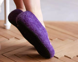 A person wearing a pair of purple felt slip-on slippers with black soles, standing on a wooden floor.