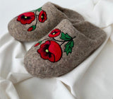 A pair of brown felt slip-on slippers with embroidered red poppy flowers and green leaves, displayed on a draped white fabric background.