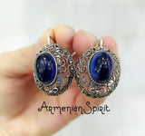 A pair of silver dome earrings with blue oval stones, held in a hand against a white background.