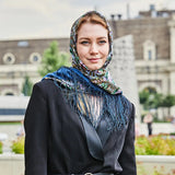 Woman wearing a patterned blue fringed headscarf and dark coat outdoors with blurred background