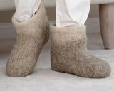 A person wearing light beige wool felt slip-on slippers like boots with white pants, standing indoors on a light floor. Authentic merino wool light gray slippers with high shift.
