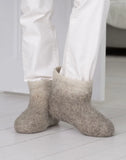 A person wearing light beige felt slipper pulled up over white pants, standing on a white wooden floor.