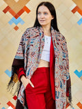 Woman wearing a colorful patterned shawl over a white top and red pants against a geometric-patterned wall. This traditional brown shawl with light blue and red ornament is inspired by Slavic folk art. A woman wearing it looks classic and refined, wrapped in soft merino wool with elegant fringe. 
