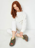 A woman with red hair sits cross-legged on a white floor against a white wall, wearing white sweatpants and a white long-sleeve top, with brown woolen winter slippers featuring green leaf trim and a red strawberry motif on her foot.
