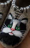A close-up of a handmade felt slipper shaped like a black and white cat's face, with green embroidered eyes, a pink nose, white whiskers, and black stitched details on a beige felt base.