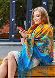A medium shot of a woman with light brown hair wearing a large, cozy merino wool shawl in a traditional Slavic style. The shawl is wrapped like a hood, showing a vibrant orange outer layer with green ornaments, while the rest of the blanket-sized fabric drapes over her beige coat in a bright blue color filled with detailed red, yellow, and green floral patterns and long matching blue fringes.