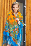 A woman smiles while wearing a warm, blanket-style winter shawl made of merino wool. The colorful garment features a striking contrast between a bright orange head covering and a brilliant blue body, both heavily decorated with traditional Slavic floral arrangements, providing a very cozy and cultural aesthetic.