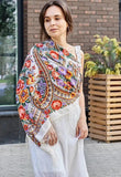 A woman wearing an oversized white merino scarf adorned with a vintage Slavic herbal pattern. The striking red and yellow details bring a retro folk charm, while the natural fabric and silky fringe add refined softness.