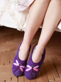 Purple felt slipper shoes with pink dragonfly embellishments, worn by a person sitting on a wooden floor next to a white blanket.