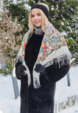 An elegant lifestyle shot featuring a blonde woman wrapped in a thick, cozy, and opaque ivory woolen shawl. The garment showcases a classic Slavic heritage design with vibrant orange and red roses, styled securely as a neck and shoulders over a black fur coat in a winter setting with blurred fairy lights.