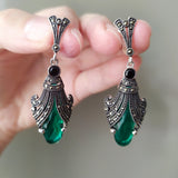 Very long silver earrings with green emerald and black marcasite stones, held in a person's hand.