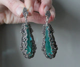 Teal earrings with silver filigree details held by a hand against a neutral background