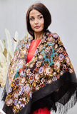 A woman wearing an authentic brown merino wool shawl full body cover enriched with beige and navy blue ornaments and delicate floral motifs. The premium merino wool feels warm and refined, while the silky fringe adds elegant movement and a luxurious finish to this timeless accessory.
