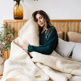 A woman sits in bed wrapped in a soft cream-colored natural merino sheepskin wool blanket, wearing a dark green top. The scene is set in a cozy bedroom with a wooden headboard, white pillows, and a glass vase with greenery on the bedside table.