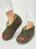 A pair of brown felt slip-on slippers with green leaf trim and embroidered red cherries, shown on a person's feet wearing white pants.