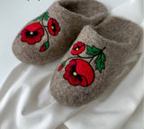 A pair of brown felted wool slip-on slippers decorated with red poppy flowers and green leaves, resting on a white satin fabric.