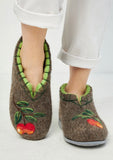 A pair of brown felt slip-on clog slippers with green leaf trim and embroidered red-orange fruit motifs, shown on a person's feet wearing white pants against a light gray background.