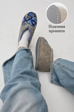 Gray warm felted wide slippers with floral design worn by a person in light blue jeans on a white background.