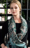 Woman wearing a green patterned woolen scarf with a blurred indoor background. This green scarf with teal and black paisley ornaments looks elegant and timeless. The ethnic, traditional design makes it easy to wear for both everyday style and special occasions. Large and comfortable.