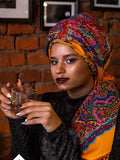 Woman wearing a colorful orange and blue headscarf holding a glass against a brick wall.